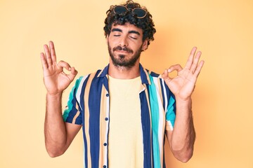 Handsome young man with curly hair and bear wearing summer shirt and sunglasses relax and smiling with eyes closed doing meditation gesture with fingers. yoga concept.