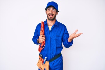 Handsome young man with curly hair and bear wearing builder jumpsuit uniform and electric cables celebrating achievement with happy smile and winner expression with raised hand