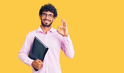 Handsome young man with curly hair and bear holding business folder doing ok sign with fingers, smiling friendly gesturing excellent symbol