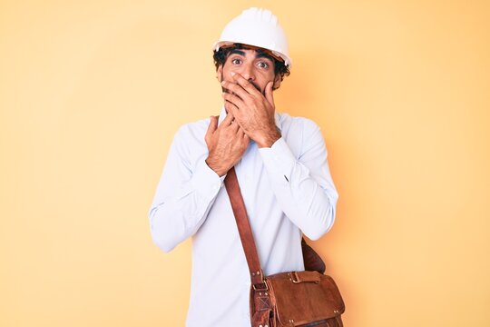 Handsome young man with curly hair and bear wearing architect hardhat shocked covering mouth with hands for mistake. secret concept.