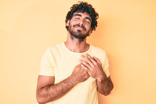 Handsome young man with curly hair and bear wearing casual tshirt over yellow background smiling with hands on chest, eyes closed with grateful gesture on face. health concept.