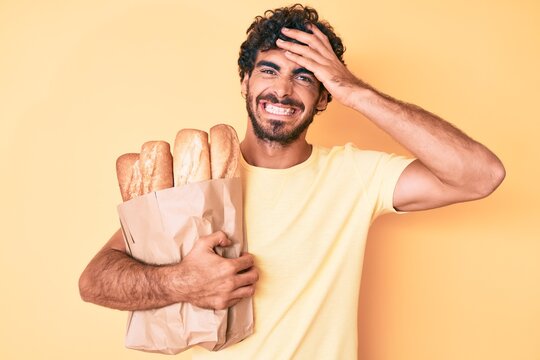 Handsome young man with curly hair and bear holding paper bag with bread stressed and frustrated with hand on head, surprised and angry face