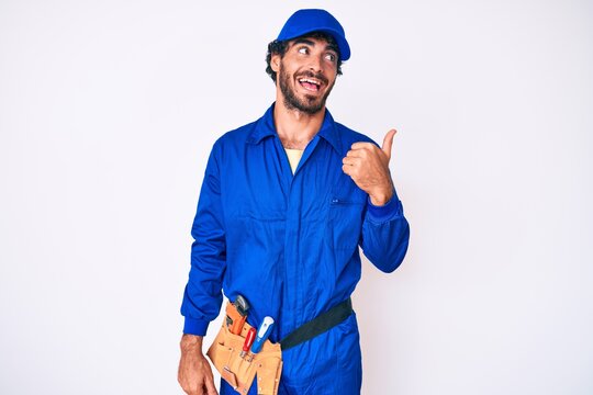 Handsome young man with curly hair and bear weaing handyman uniform smiling with happy face looking and pointing to the side with thumb up.