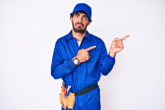 Handsome young man with curly hair and bear weaing handyman uniform pointing aside worried and nervous with both hands, concerned and surprised expression