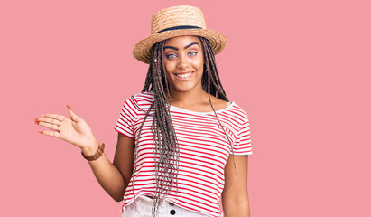 Obraz na płótnie Canvas Young african american woman with braids wearing summer hat smiling cheerful presenting and pointing with palm of hand looking at the camera.