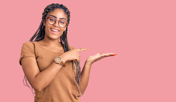 Young african american woman with braids wearing casual clothes and glasses amazed and smiling to the camera while presenting with hand and pointing with finger.