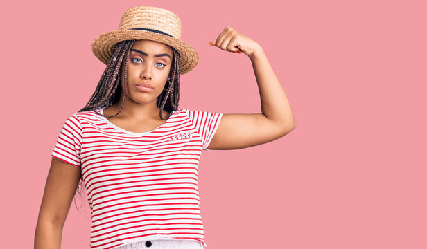 Young african american woman with braids wearing summer hat strong person showing arm muscle, confident and proud of power
