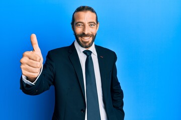 Attractive man with long hair and beard wearing business suit and tie smiling happy and positive, thumb up doing excellent and approval sign