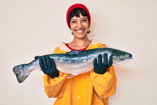 Beautiful brunettte fisher woman wearing raincoat holding fresh salmon smiling with a happy and cool smile on face. showing teeth.