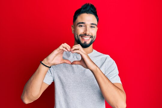 Young man with beard wearing casual white tshirt smiling in love doing heart symbol shape with hands. romantic concept.
