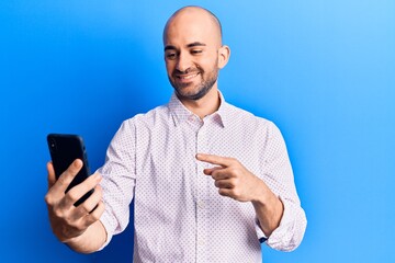 Young handsome bald man using smartphone smiling happy pointing with hand and finger