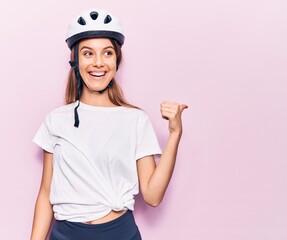 Young beautiful girl wearing bike helmet pointing thumb up to the side smiling happy with open mouth