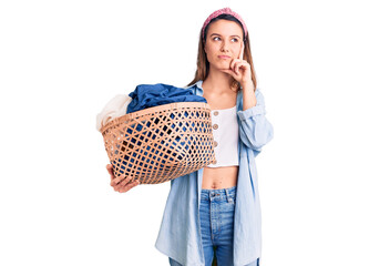 Young beautiful girl holding laundry basket serious face thinking about question with hand on chin,...