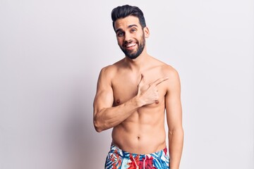 Young handsome man with beard shirtless wearing swimwear smiling cheerful pointing with hand and finger up to the side