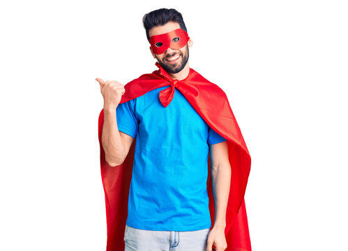 Young handsome man with beard wearing super hero costume smiling with happy face looking and pointing to the side with thumb up.