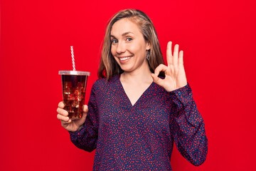 Young beautiful blonde woman drinking a fresh glass of soda doing ok sign with fingers, smiling friendly gesturing excellent symbol