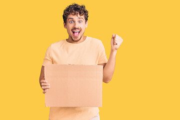 Young handsome man with curly hair holding blank cardboard empty banner screaming proud, celebrating victory and success very excited with raised arms