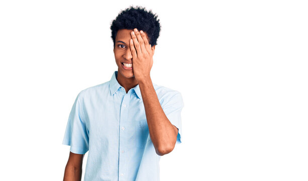 Young african american man wearing casual clothes covering one eye with hand, confident smile on face and surprise emotion.