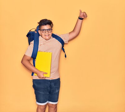Adorable student boy wearing glasses and backpack smiling happy. Jumping with smile on face holding book doing ok sign with thumb up over isolated yellow background.