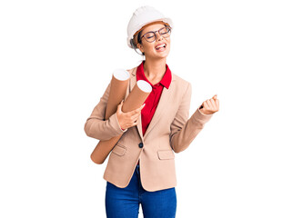 Young beautiful woman wearing architect hardhat and glasses holding blueprints screaming proud, celebrating victory and success very excited with raised arms