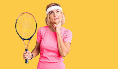 Young blonde woman playing tennis holding racket serious face thinking about question with hand on chin, thoughtful about confusing idea
