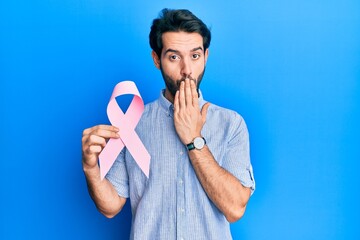 Young hispanic man holding pink cancer ribbon covering mouth with hand, shocked and afraid for mistake. surprised expression