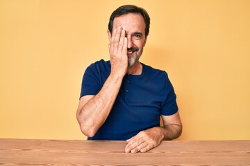 Middle age hispanic man wearing casual clothes sitting on the table covering one eye with hand, confident smile on face and surprise emotion.