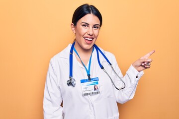 Young beautiful doctor woman wearing stethoscope and id card over isolated yellow background smiling cheerful pointing with hand and finger up to the side