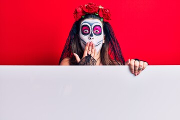 Young woman wearing day of the dead costume holding blank empty banner covering mouth with hand, shocked and afraid for mistake. surprised expression