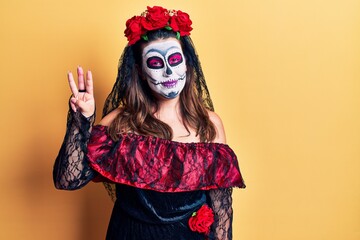 Young woman wearing day of the dead costume over yellow showing and pointing up with fingers number three while smiling confident and happy.