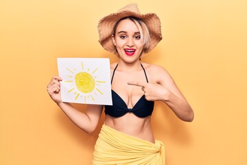 Young beautiful blonde woman wearing bikini and summer hat holding sun draw smiling happy pointing with hand and finger