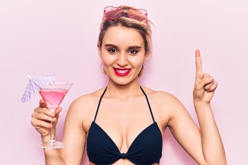 Young beautiful blonde woman wearing bikini drinking cocktail smiling with an idea or question pointing finger with happy face, number one