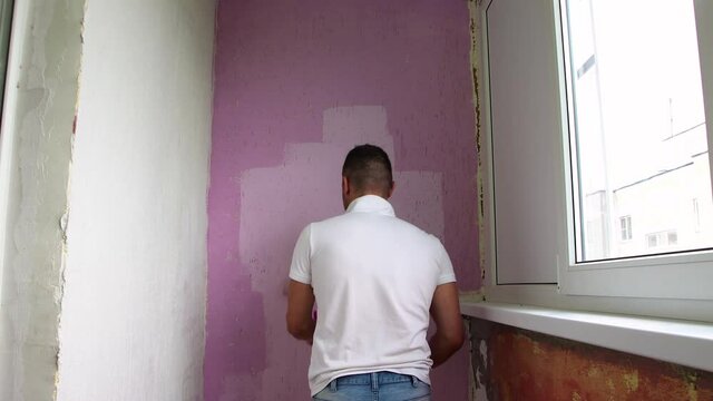 A young man with his back turned in a white T-shirt is painting the wall. The master does the work with a paint roller. It covers the surface with pink paint. Daylight.