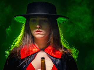 Vape device in hand. Close-up portrait of a woman vaper. Vaper girl in hat. Young girl holding a vape pen. Girl with electronic cigarette in neon light. Woman with a vape device on a green background