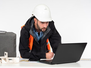 Construction concept on a white background. A civil engineer looks at the details of the project on his laptop. A man in a construction helmet at a table with a layout of the house and drawings.