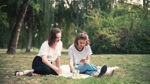 Two young cheerful women sitting on a blanket in the park looking an the phone and drinking wine while on a picnic.