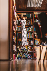 Full length of young attractive librarian standing on ladder and searching for a book.