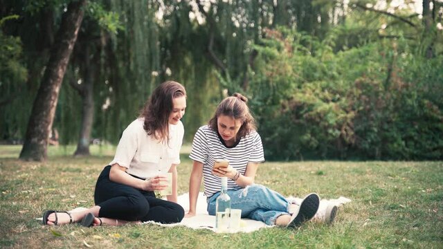 Two young women are making a picnic with wine. Friends are sitting on a blanket in the park, looking at the phone and talking.