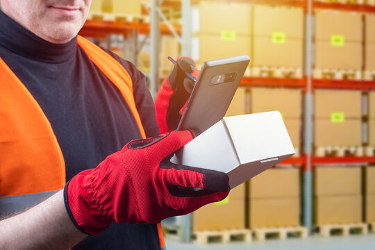 A man with a cardboard box and a smartphone in his hands.The person makes a note in the smartphone about the availability of the product in stock. Storekeeper with a smartphone. Working in a warehouse