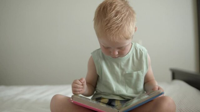 Little toddler boy reading book at home, turning pages, looking at the colorful pictures, home education, kids books.