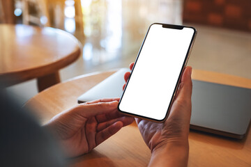 Mockup image of a woman holding mobile phone with blank white desktop screen with laptop on the table