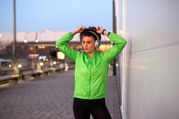 Fit woman taking a break and standing with headphones over her head after jogging at street at night