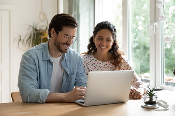Smiling young Caucasian couple sit at desk at home look at laptop screen shopping online together. Happy husband and wife browse wireless internet on computer. Technology, communication concept.