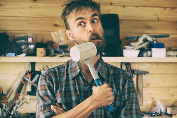Funny bearded worker man stands in workshop and hits himself with a mallet, construction carpentry...