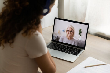 Back view of young woman have webcam conference conversation with smiling elderly mother. Female sit at desk at home talk on video call online with teacher or trainer. Distant communication concept.