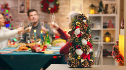 Little christmas tree on focus with family celebrating christmas in the background. Traditional festive christmas dinner in multigenerational family. Enjoying xmas meal feast in decorated room. Big