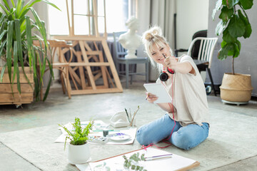 Happy laughing girl artist, painter, designer working at studio workshop. Positive overjoyed young woman in casual clothes and headphones showing pencil to camera sitting on floor. Creative hobby