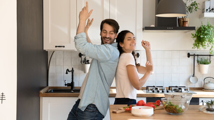 Overjoyed young couple renters have fun in kitchen celebrating moving to new home together. Happy millennial man and woman tenants dance near counter cooking healthy food. Relocation concept.