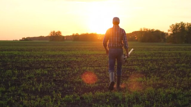 Young male farmer man in rubber boots, shirt holding shovel on vegetables growing field of wheat at sunset spring. Farming Agriculture harvesting food farmland ground concept, close-up back view, 4 K