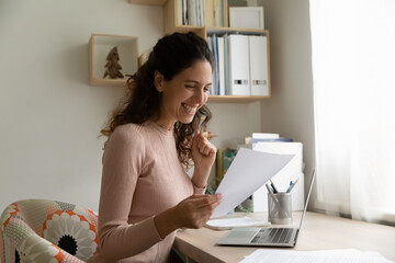 Overjoyed millennial Caucasian woman work on laptop at home triumph reading good news in paperwork...